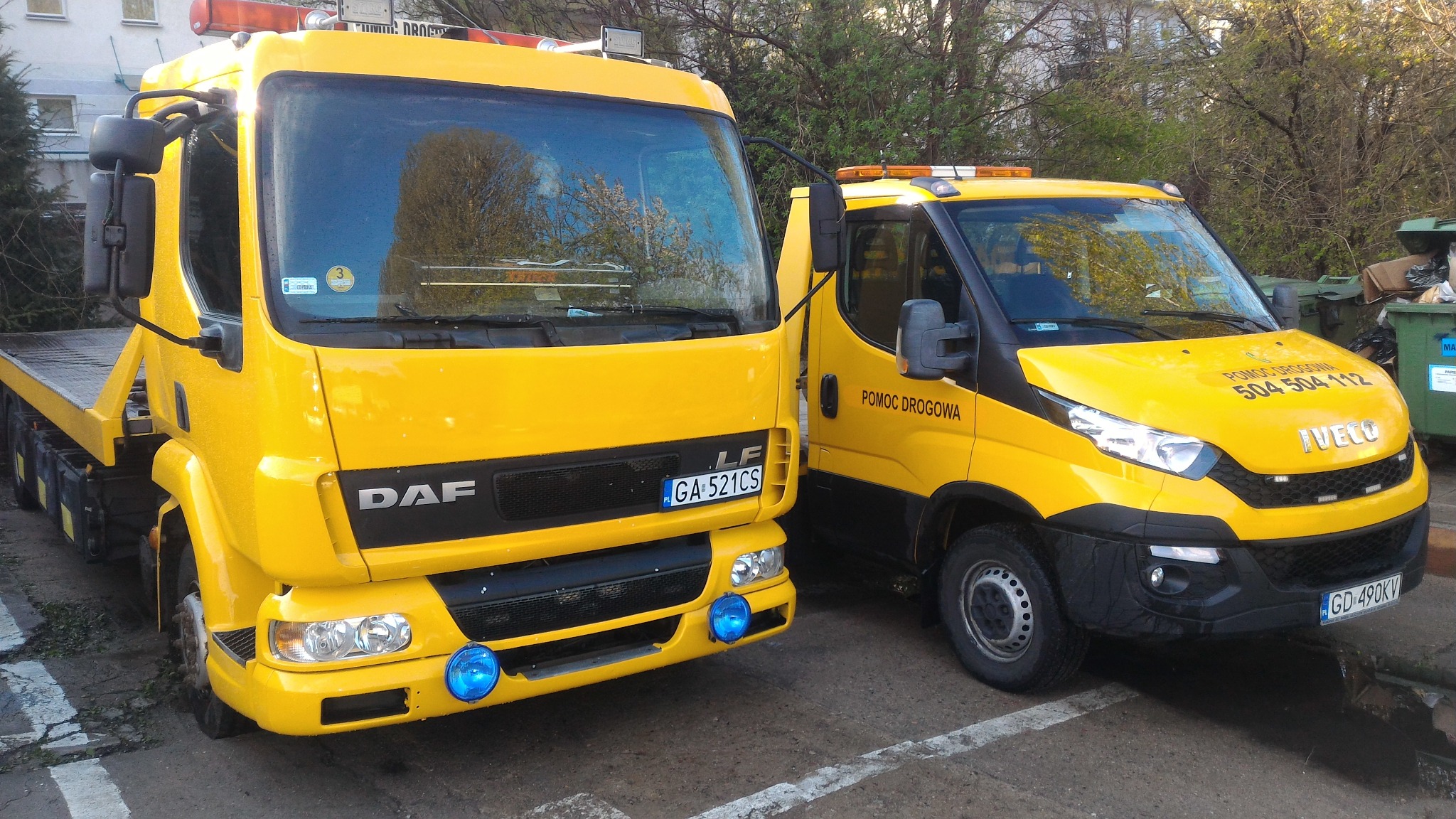 Dwa żółte pojazdy pomocy drogowej: DAF laweta z niebieskimi światłami i Iveco z numerem telefonu, stojące na parkingu.