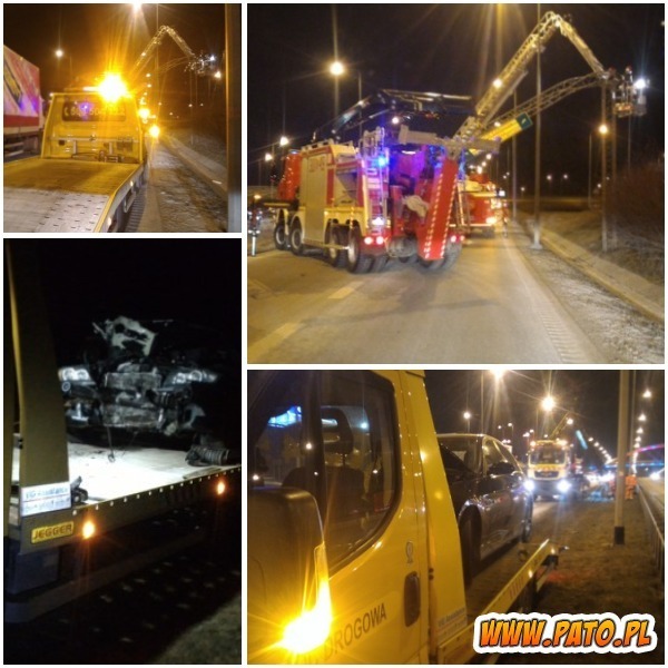 Nocne działanie służb ratunkowych na drodze: laweta z uszkodzonym pojazdem, w tle wóz strażacki z podnośnikiem.