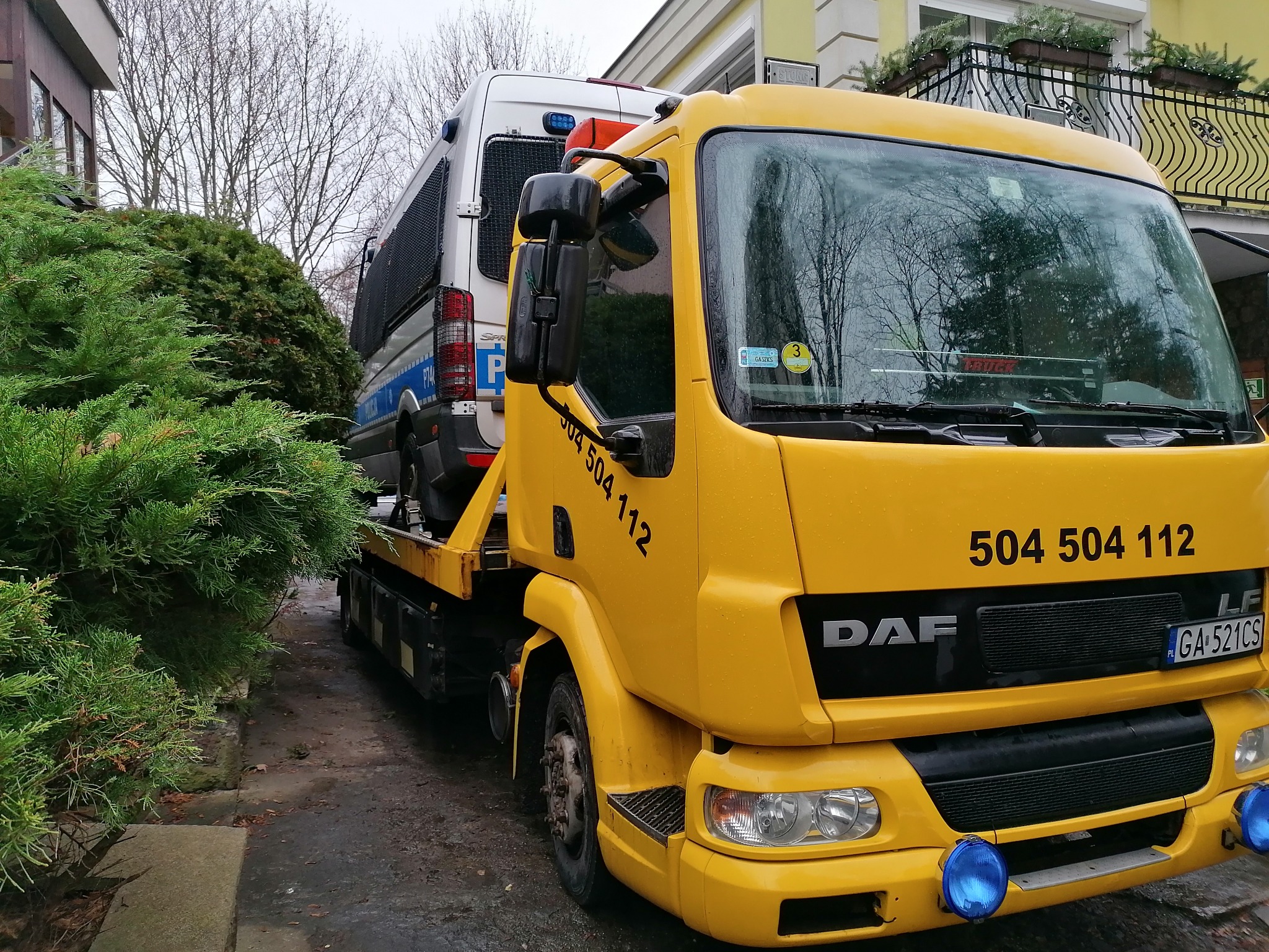 Żółta laweta DAF z numerem telefonu 504 504 112 przewozi policyjny samochód dostawczy na tle budynku mieszkalnego z balkonem i zielenią.