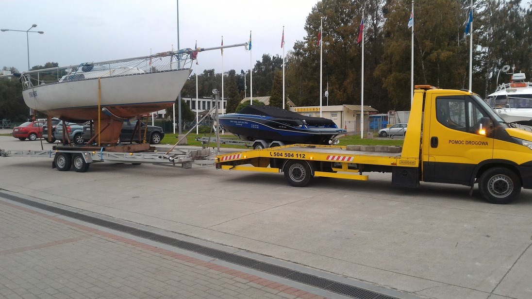 Żółta laweta 'Pomoc Drogowa' transportuje jacht żaglowy na przyczepie, obok stoi przykryta plandeką motorówka na drugiej przyczepie, w tle maszty z flagami.