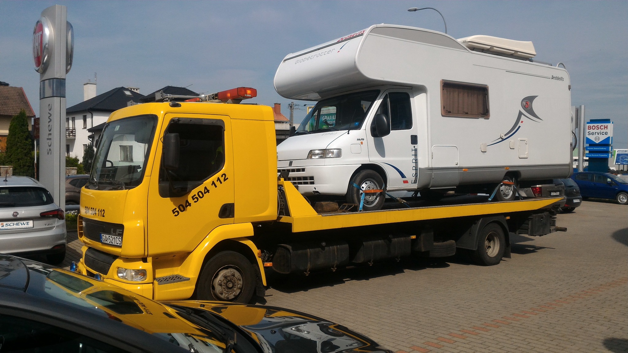 Żółta ciężarówka pomocy drogowej z numerem telefonu 504 504 112 przewozi biały kamper marki Dethleffs na platformie. Kamper zabezpieczony pasami transportowymi. W tle widoczny salon Fiata i znak...