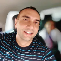 Uśmiechnięty mężczyzna w samochodzie we Wrocławiu, ubrany w koszulkę w paski, z rozmazaną postacią w tle.