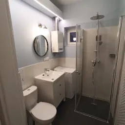 Jasna łazienka po remoncie z kabiną prysznicową typu walk-in, umywalką z szafką, toaletą, pralką i okrągłym lustrem na ścianie.