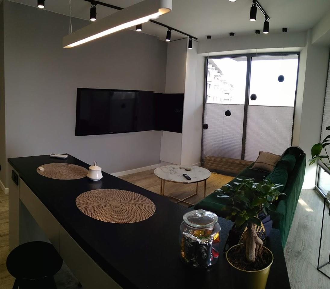 Nowoczesny salon z aneksem kuchennym w odcieniach szarości, czerni i zieleni. Widoczne: telewizor na ścianie, wyspa kuchenna z dekoracyjnymi podkładkami, okrągły stolik kawowy i zielona sofa.