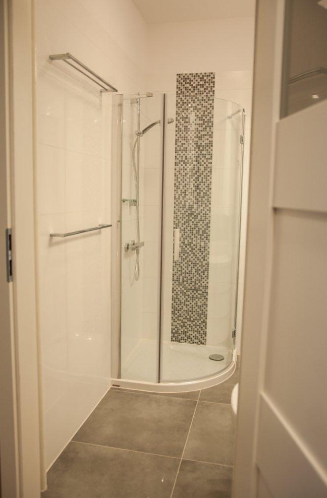 Nowoczesna kabina prysznicowa z mozaiką w wyremontowanej łazience. Szklane drzwi, białe płytki i szare podłogowe. Widok zza otwartych drzwi.