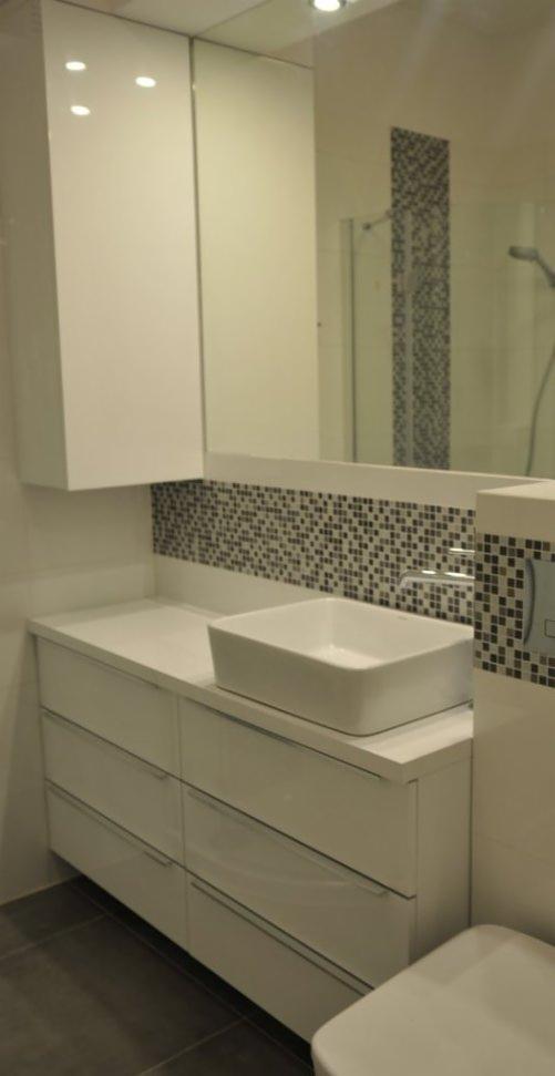 Biała, nowoczesna łazienka z szafkami pod umywalką, lustrem i mozaiką. Minimalistyczny design, prostokątna umywalka nablatowa, szare płytki podłogowe.