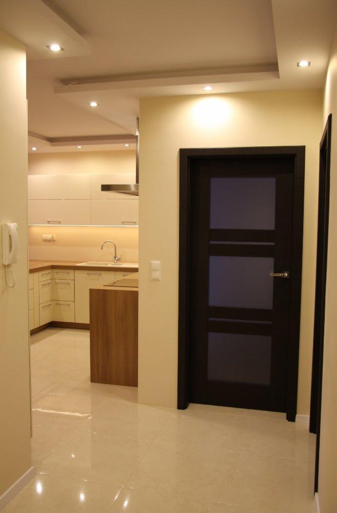 Jasne wnętrze mieszkania po remoncie: widok na kuchnię z wyspą i ciemne drzwi z przeszkleniami. Beżowe ściany, błyszcząca podłoga, nowoczesne oświetlenie.