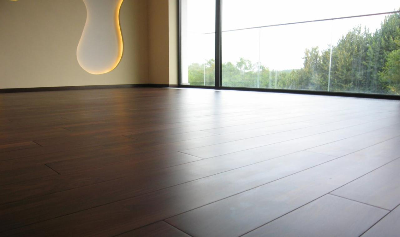 Parkiet z ciemnego drewna w jasnym pomieszczeniu z dużym oknem i minimalistyczną lampą ścienną. Widok na zieleń za oknem.