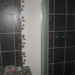 Wykończona kabina prysznicowa z czarno-białymi płytkami zdobionymi motywem kwiatowym. Widoczne narzędzia na blacie sugerują etap prac wykończeniowych.