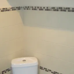 Jasna łazienka po remoncie: beżowe płytki, dekoracyjny pasek z mozaiki, toaleta i umywalka. Widoczne gniazdko i wieszak na ręczniki.