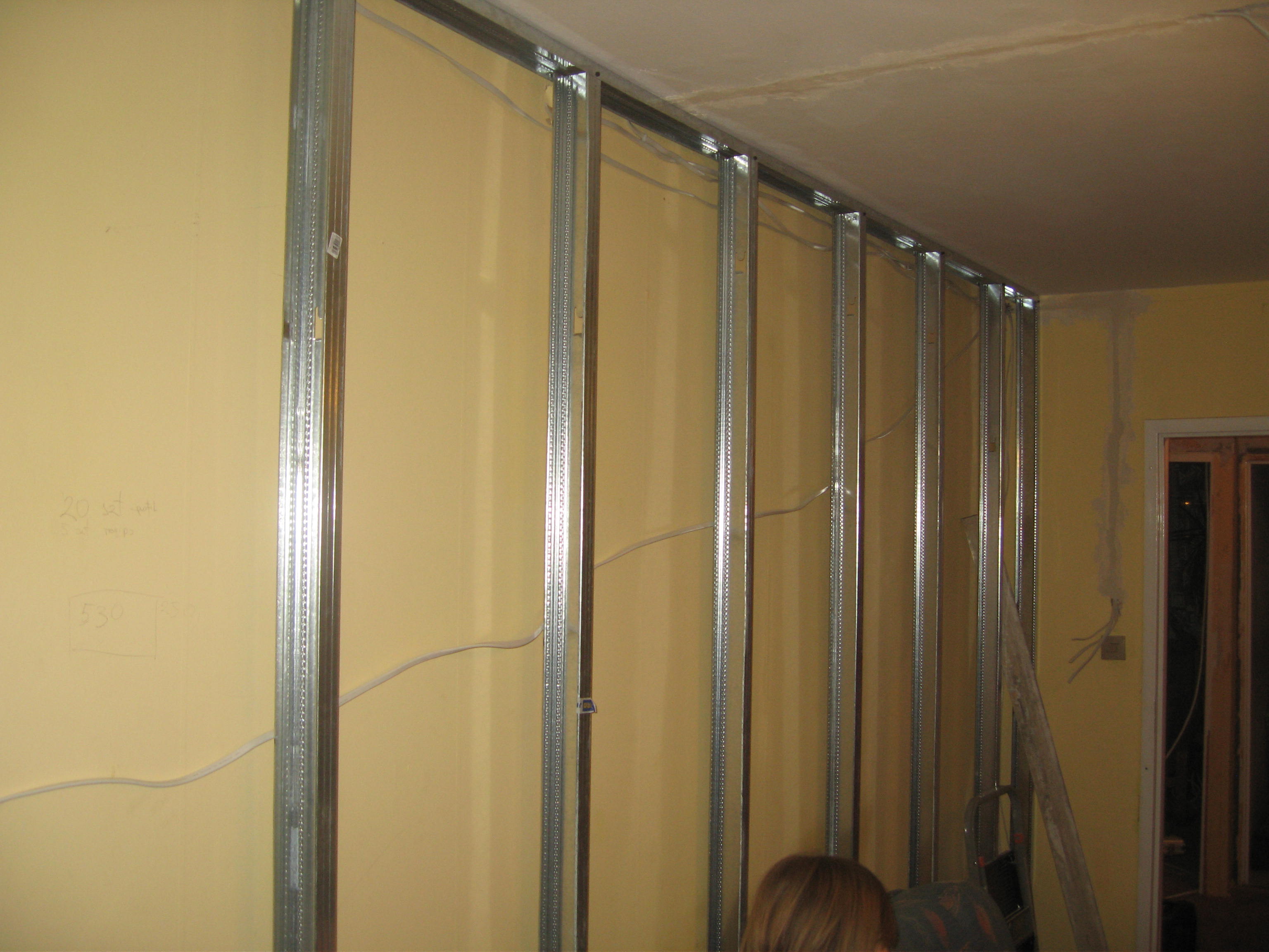 Konstrukcja ścianki działowej z metalowych profili w trakcie montażu. Widoczne przewody elektryczne i żółta ściana. W tle fragment drzwi.