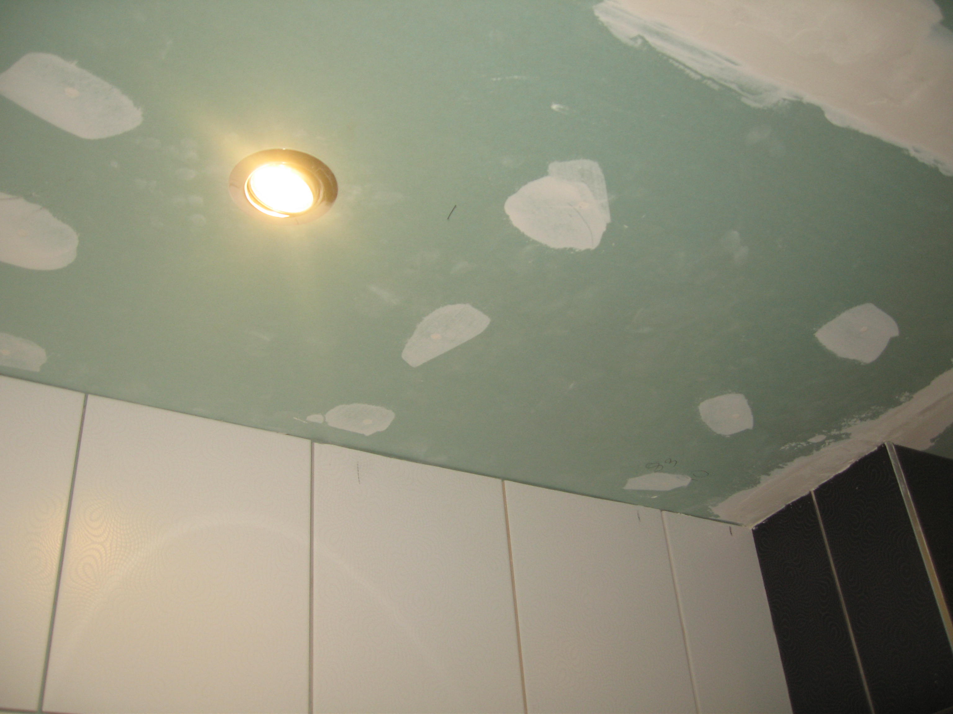 Zielonkawy sufit z zamontowanym oświetleniem punktowym i białymi plamami po szpachlowaniu, częściowo widoczne białe i czarne płytki na ścianie.