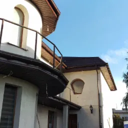 Kremowa elewacja domu z brązowymi elementami wykończeniowymi i nietypowym oknem w kształcie trapezu, widok z dołu na błękitne niebo i drzewo.