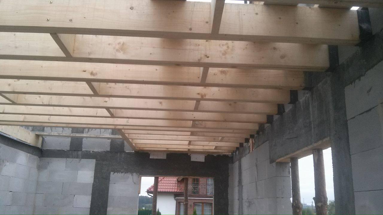 Surowy stan budowy domu z bloczków, widoczne drewniane belki stropowe i fragment dachu sąsiedniego budynku przez otwór okienny.