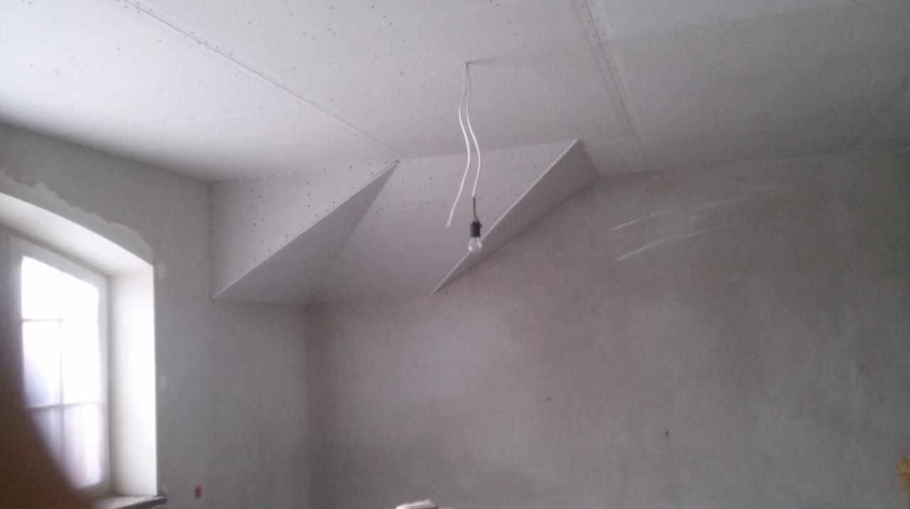 Wnętrze pokoju w trakcie remontu: sufit z płyt gipsowo-kartonowych ze skośnymi elementami, surowe ściany, wisząca żarówka i okno z łukiem.
