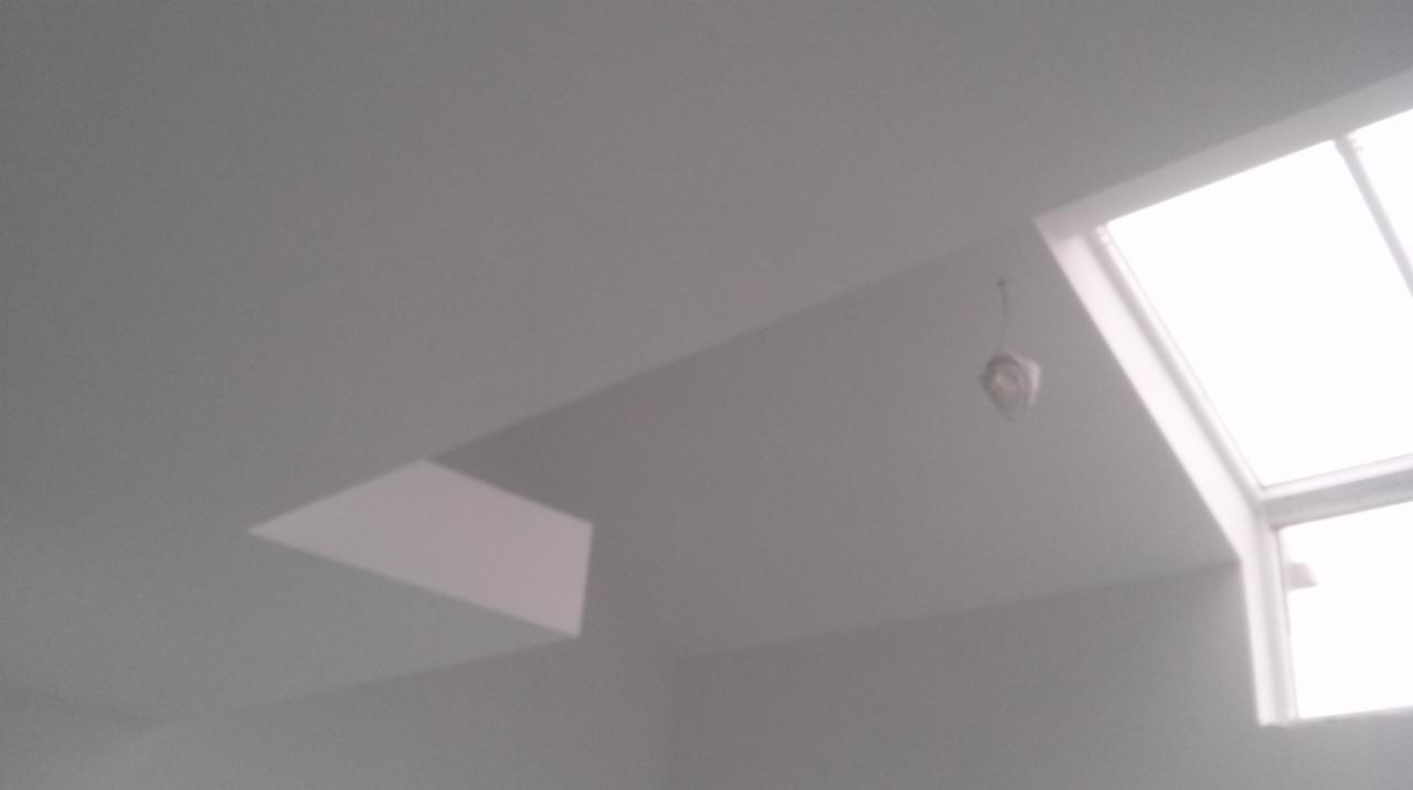 Jasne, świeżo pomalowane wnętrze z oknem dachowym i charakterystycznym załamaniem sufitu. Widoczny fragment instalacji oświetleniowej.