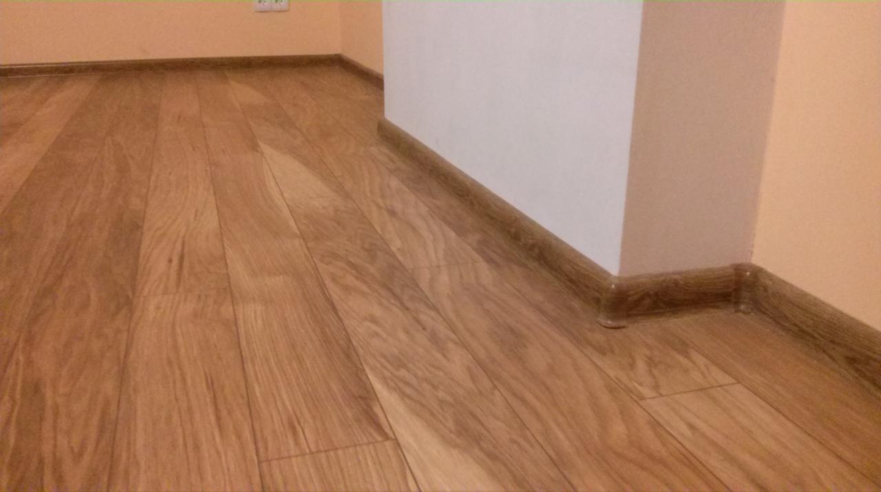 Ułożona podłoga z paneli z drewnianym wzorem, widoczne listwy przypodłogowe i fragment ściany w jasnych odcieniach. Ujęcie z niskiego kąta.