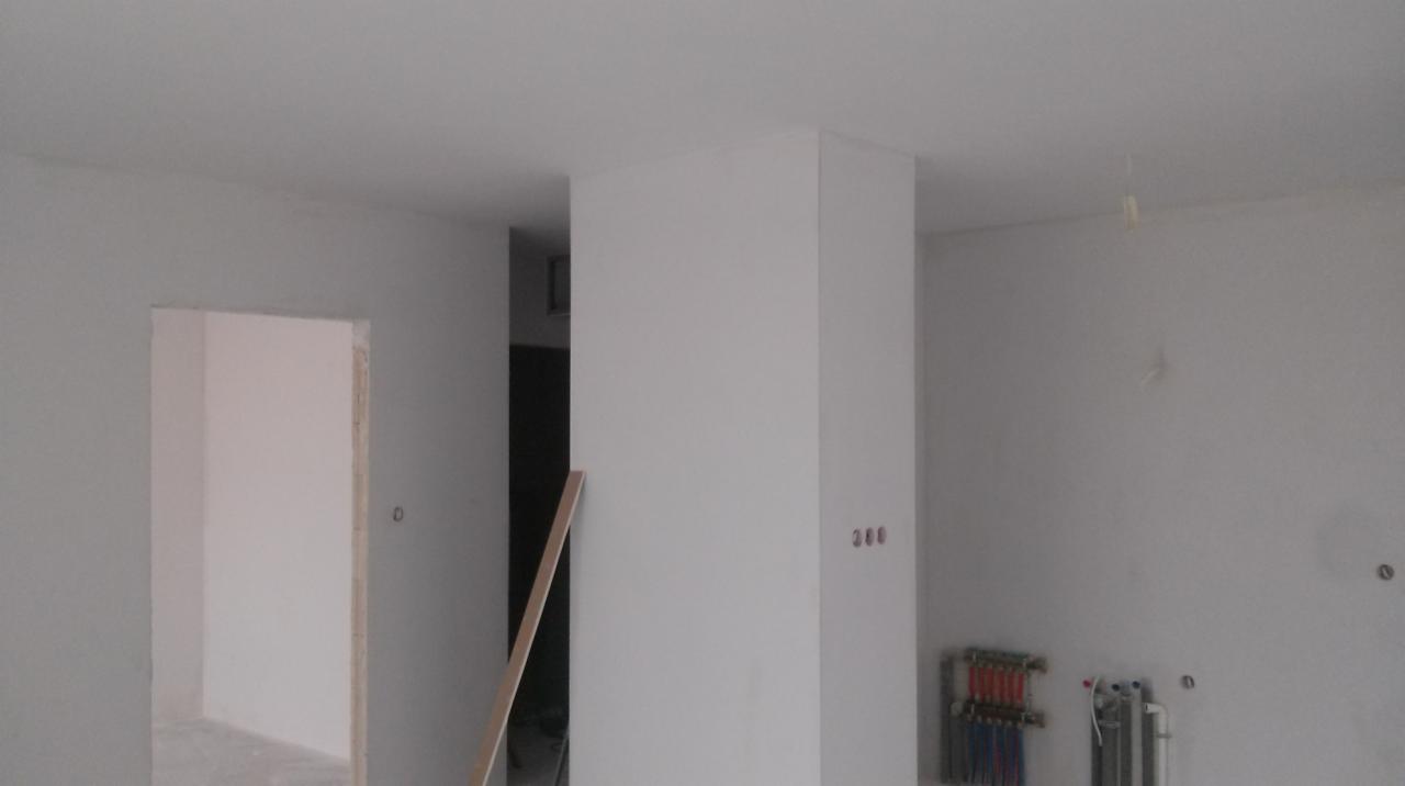 Wnętrze w trakcie wykończenia: białe ściany, widoczne instalacje grzewcze, surowe przejście w ścianie. Prace wykończeniowe w nowym budownictwie.