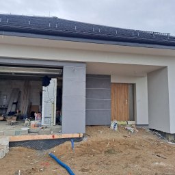 EXPOL Docieplenia budynków Jakub Brzeski - Nowo wybudowany dom jednorodzinny w trakcie prac wykończeniowych elewacji, z widocznymi elementami ocieplenia fundamentów, surowym wnętrzem garażu i częściowo otynkowaną fasadą w odcieniach...