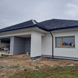 EXPOL Docieplenia budynków Jakub Brzeski - Nowoczesny dom jednorodzinny z jasną elewacją i ciemną dachówką w trakcie prac wykończeniowych, widoczny fragment izolacji fundamentów oraz otwarty garaż z szarymi ścianami.