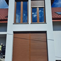 EXPOL Docieplenia budynków Jakub Brzeski - Dwukondygnacyjny fragment elewacji domu z zamontowanymi brązowymi roletami zewnętrznymi, częściowo opuszczonymi. W oknach odbija się błękitne niebo. Na dole widoczna izolacja fundamentów. W tle...