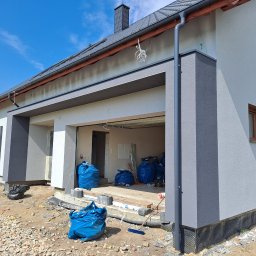 EXPOL Docieplenia budynków Jakub Brzeski - Nowoczesny dom jednorodzinny w trakcie prac elewacyjnych i izolacji fundamentów, widoczne worki z materiałami budowlanymi i narzędzia.