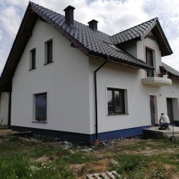 EXPOL Docieplenia budynków Jakub Brzeski - Nowy dom z białą elewacją, czarną dachówką i pracownikami wykańczającymi detale wokół okien i na schodach wejściowych, widoczny kontener na gruz.