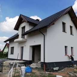 EXPOL Docieplenia budynków Jakub Brzeski - Dwukondygnacyjny dom jednorodzinny z białymi ścianami elewacyjnymi w trakcie wykańczania, widoczny pracownik malujący obramowanie okna na pomarańczowo, czarna podmurówka, dach z ciemnej dachówki...