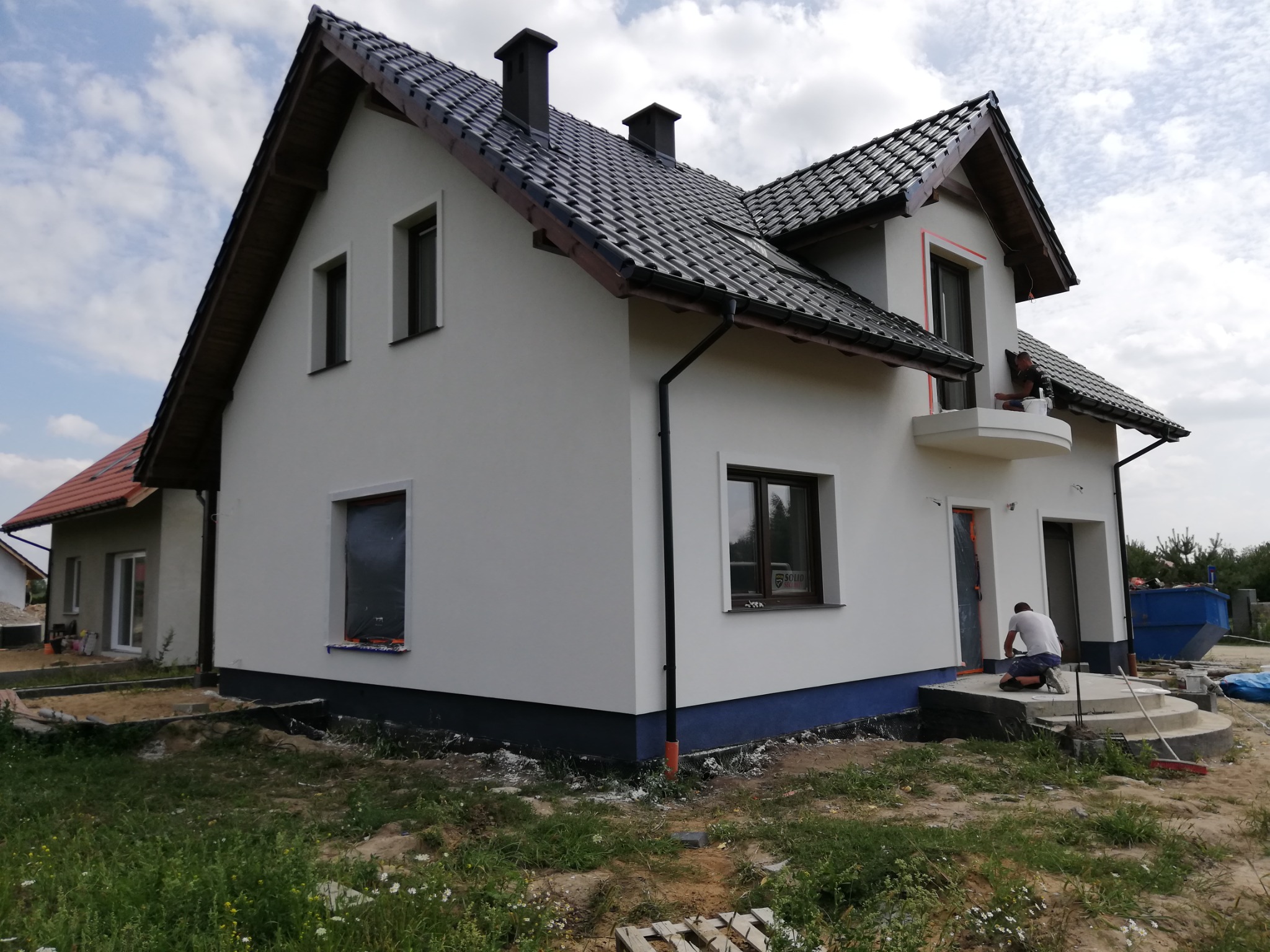 Nowy dom z białą elewacją, czarną dachówką i pracownikami wykańczającymi detale wokół okien i na schodach wejściowych, widoczny kontener na gruz.