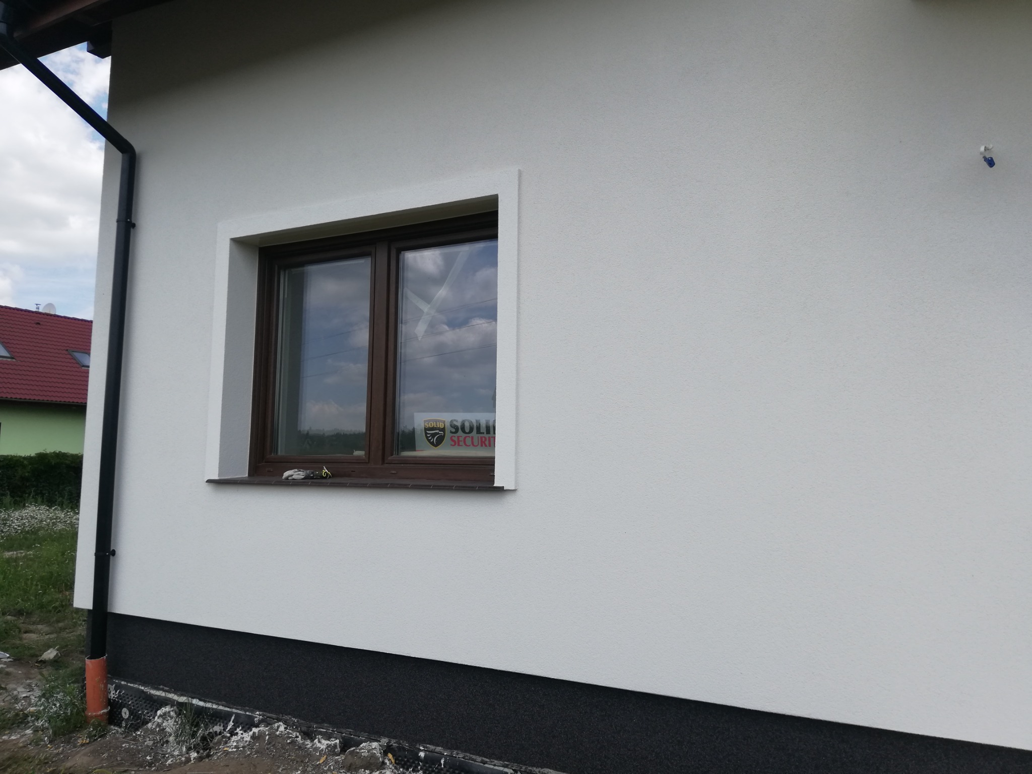 Fragment ściany domu z oknem i nową, jasną elewacją, widoczny fragment rynny i cokół z ciemnym wykończeniem, na szybie naklejka firmy ochroniarskiej.