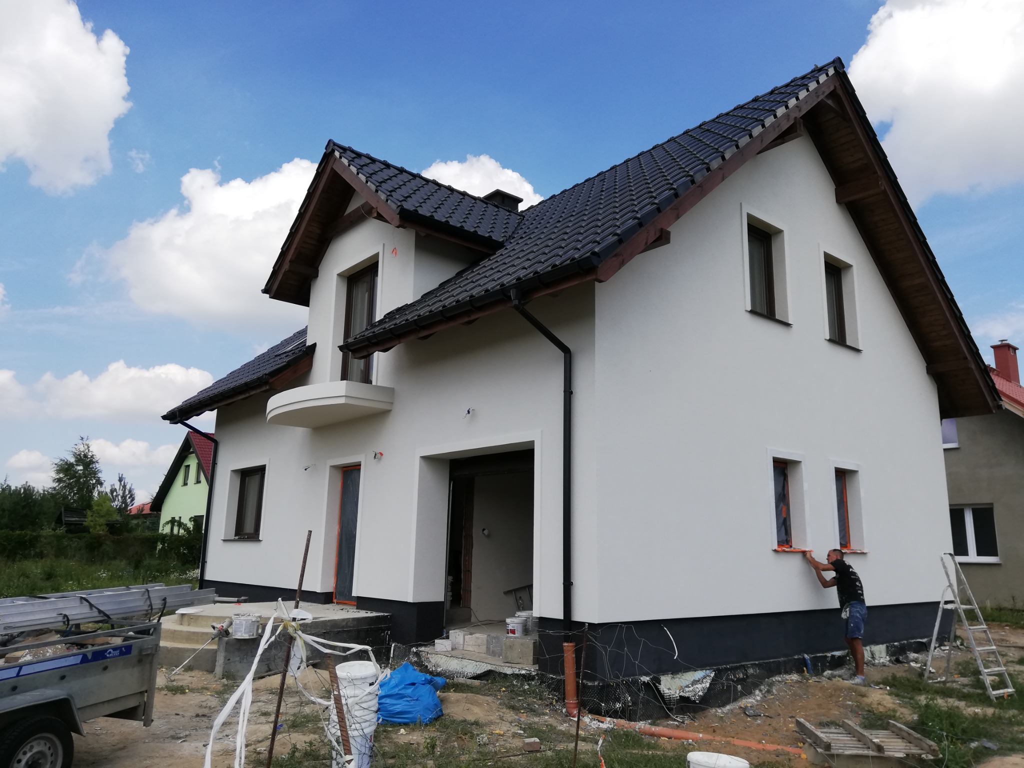 Dwukondygnacyjny dom jednorodzinny z białymi ścianami elewacyjnymi w trakcie wykańczania, widoczny pracownik malujący obramowanie okna na pomarańczowo, czarna podmurówka, dach z ciemnej dachówki...