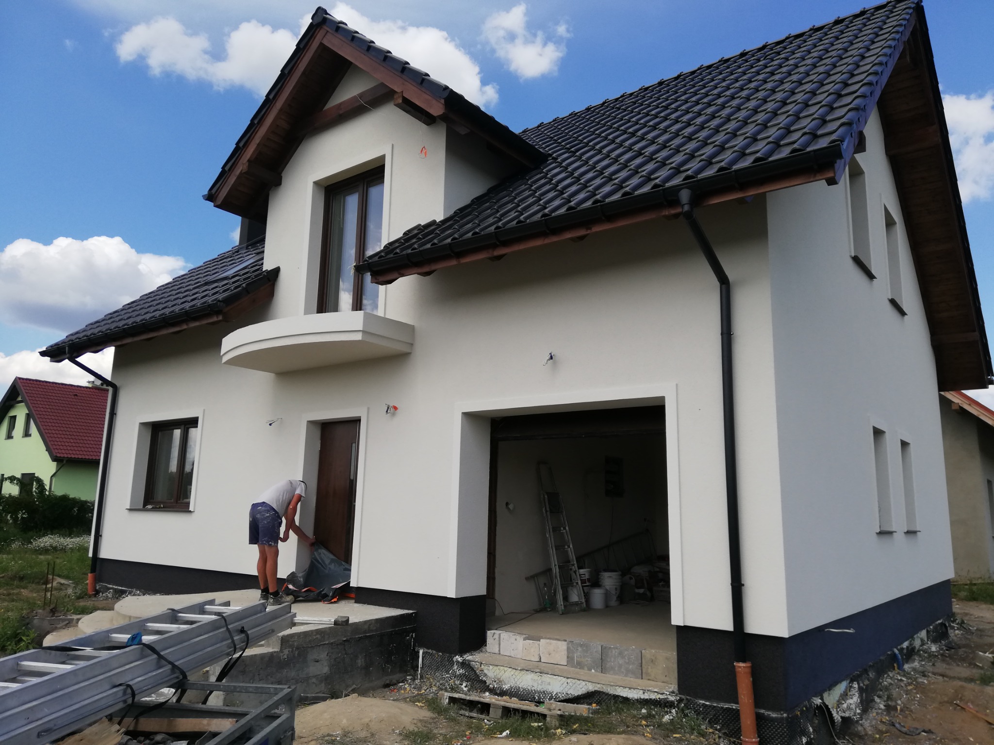 Nowo otynkowany dom jednorodzinny z ciemną dachówką i drewnianymi elementami wykończeniowymi; pracownik przygotowuje wejście do zabezpieczenia przed zabrudzeniem podczas dalszych prac.