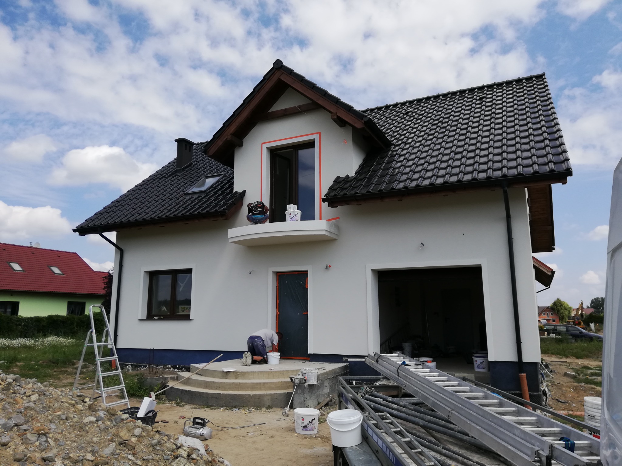 Dwukondygnacyjny dom jednorodzinny z szarą dachówką, przygotowany do tynkowania elewacji, z widocznymi zabezpieczeniami okien i drzwi, oraz pracownikiem przy wejściu, z drabina i sprzętem...