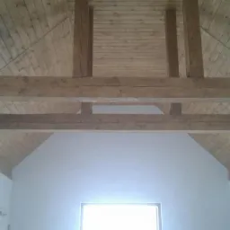 Drewniana konstrukcja stropu z belkami w jasnym pomieszczeniu z oknami. Widok z dołu na sufit ze skośnym dachem i widocznymi elementami więźby dachowej.