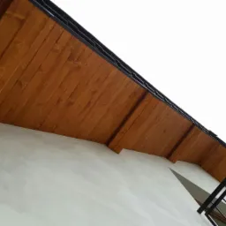 Ściana budynku po nałożeniu szarego tynku, z widocznym drewnianym podbitkiem dachowym w kolorze brązowym i fragmentem metalowej balustrady balkonowej.