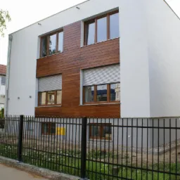 Nowoczesny, dwupiętrowy dom z białymi ścianami i drewnianą elewacją. Okna z brązowymi ramami, częściowo zasłonięte roletami. Czarny metalowy płot na pierwszym planie.