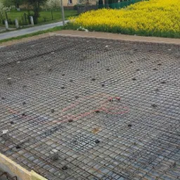 Zbrojenie płyty fundamentowej z widoczną siatką stalową, dystansami i częściowo wykonanym szalunkiem. W tle budynek i pole rzepaku.