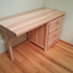 Biurko z jasnego drewna z czterema szufladami i ozdobnymi uchwytami, ustawione na parkiecie w rogu pomieszczenia z jasnozielonymi ścianami.