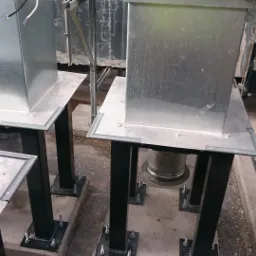 Montaż kanałów wentylacyjnych: metalowe, kwadratowe kanały na czarnych, stalowych podporach, widoczne śruby mocujące i fragment instalacji. Czerwona skrzynka narzędziowa Hilti w tle.