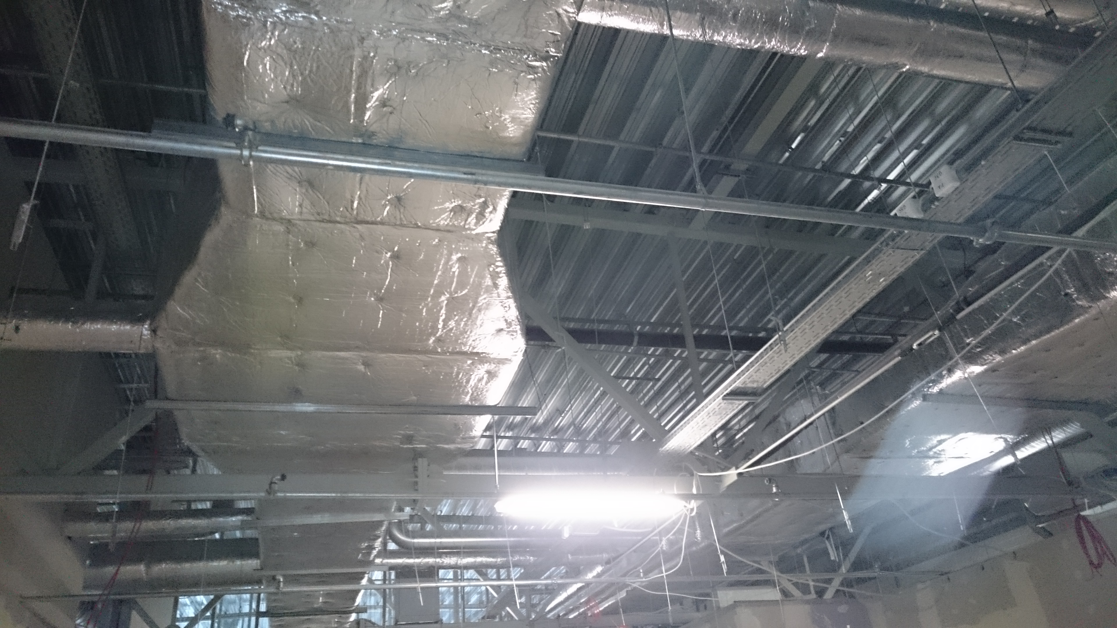 Widok instalacji pod sufitem: rury owinięte folią izolacyjną, metalowe konstrukcje i kable. Oświetlenie jarzeniowe. Całość sprawia wrażenie surowego stanu wykończenia.