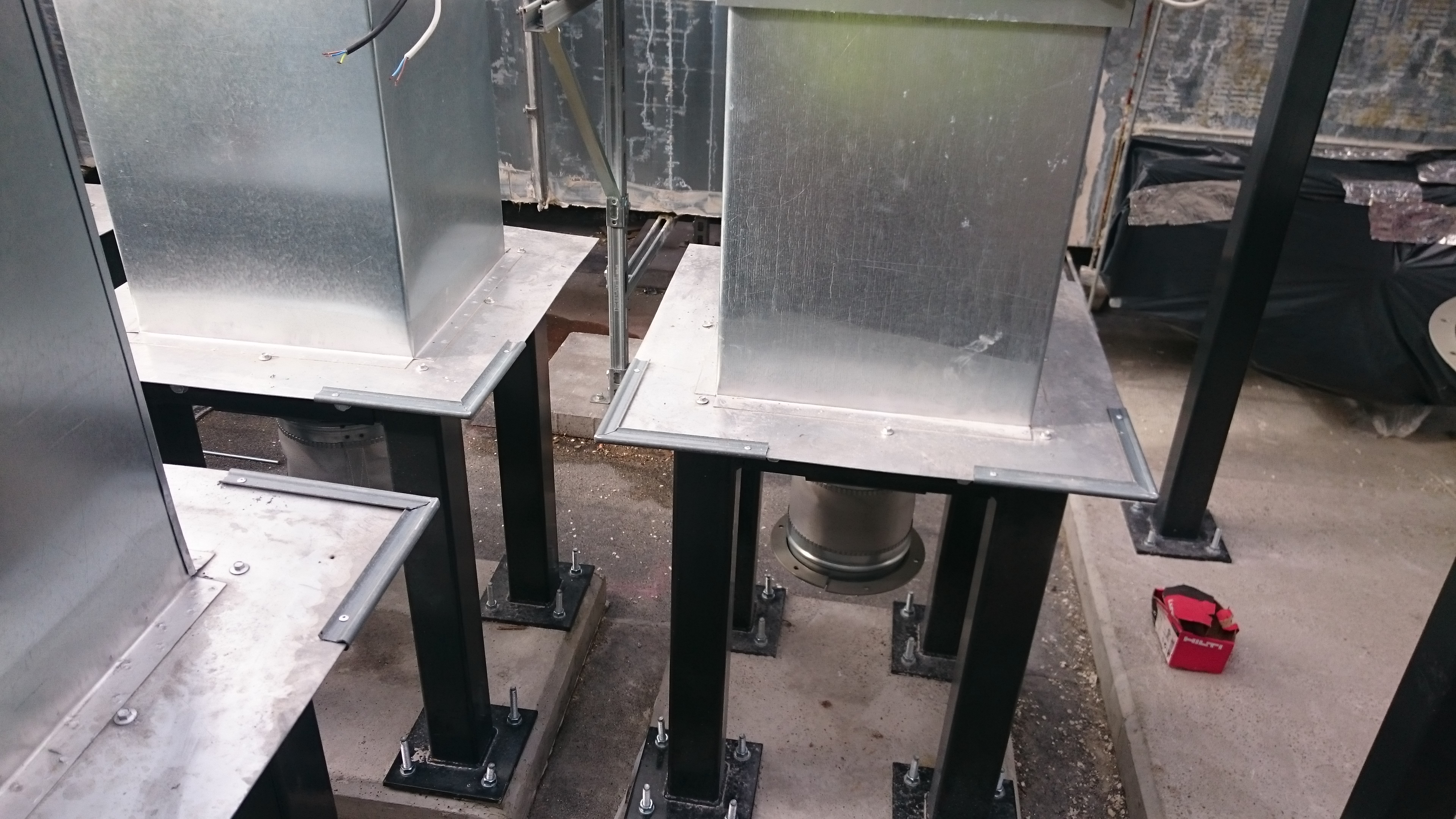 Montaż kanałów wentylacyjnych: metalowe, kwadratowe kanały na czarnych, stalowych podporach, widoczne śruby mocujące i fragment instalacji. Czerwona skrzynka narzędziowa Hilti w tle.