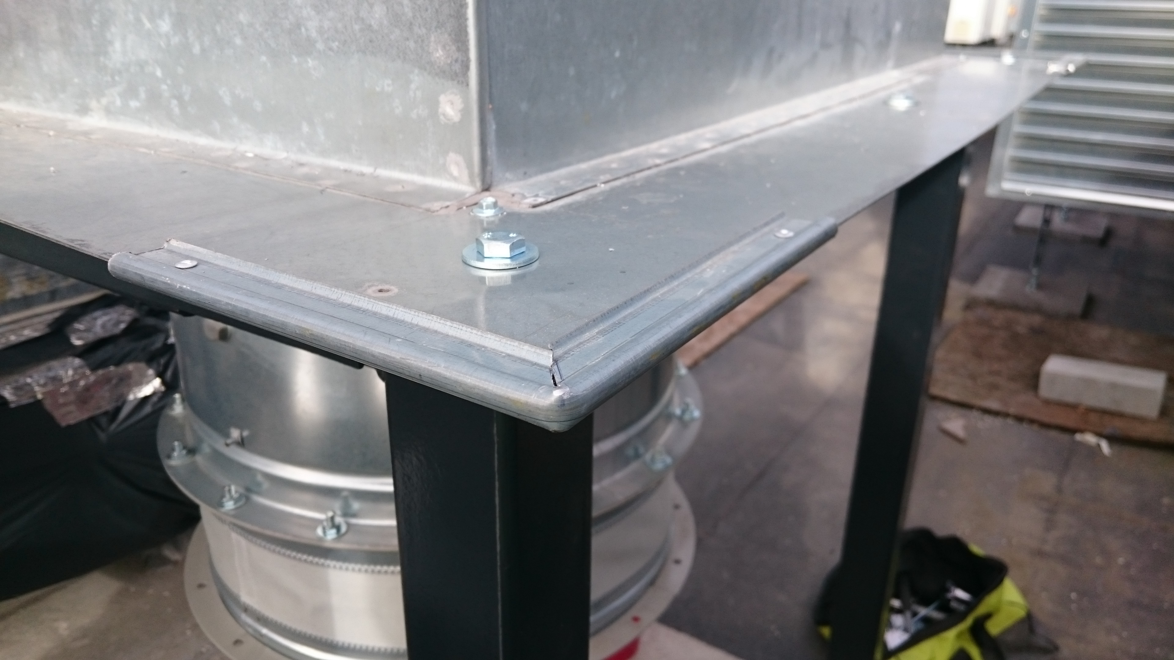 Szczegółowe ujęcie metalowej konstrukcji wentylacyjnej z elementami mocującymi, widoczne fragmenty instalacji i podłoża.