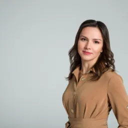 Portret uśmiechniętej kobiety w brązowej sukience koszulowej z ozdobnymi złotymi guzikami na jednolitym szarym tle.