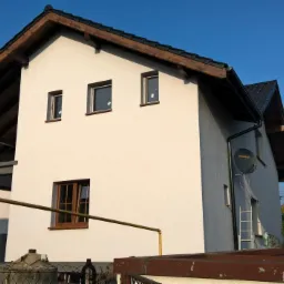 Dwukondygnacyjny dom z jasną elewacją, drewnianym dachem i brązowymi oknami. Widoczna drabina oparta o ścianę i instalacja gazowa. Błękitne niebo w tle.