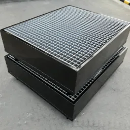 Dwa czarne, metalowe zbiorniki z kratownicą, ustawione jeden na drugim na betonowej posadzce w hali.