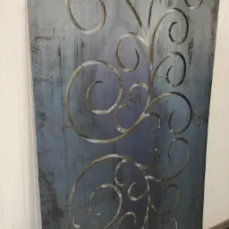 Dekoracyjny panel metalowy z wyciętym wzorem roślinnym na szarym tle z efektem rdzy, widoczne fragmenty białej ściany.