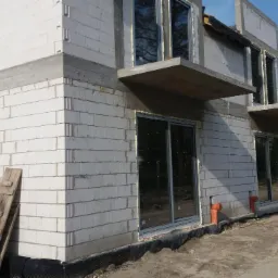 Budowa osiedla Złoty Jasieniec: surowy stan z zamontowanymi oknami przesuwnymi, widoczne bloczki z betonu komórkowego, izolacja fundamentów i prowizoryczne drewniane schody.