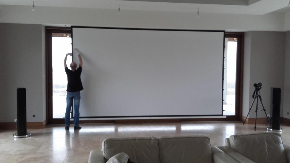 Instalacja dużego ekranu projekcyjnego w jasnym salonie, widoczne głośniki podłogowe i kamera na statywie, mężczyzna w rękawicach dopasowuje górną krawędź ekranu.