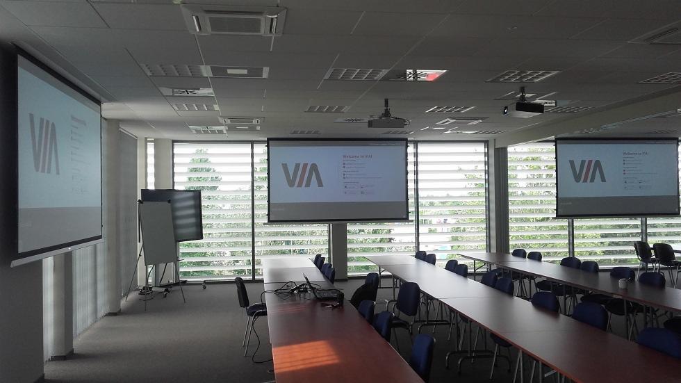 Sala konferencyjna z dwoma ekranami projekcyjnymi wyświetlającymi logo VIA, rzędy stołów i krzeseł, oraz projektorami zamontowanymi na suficie.