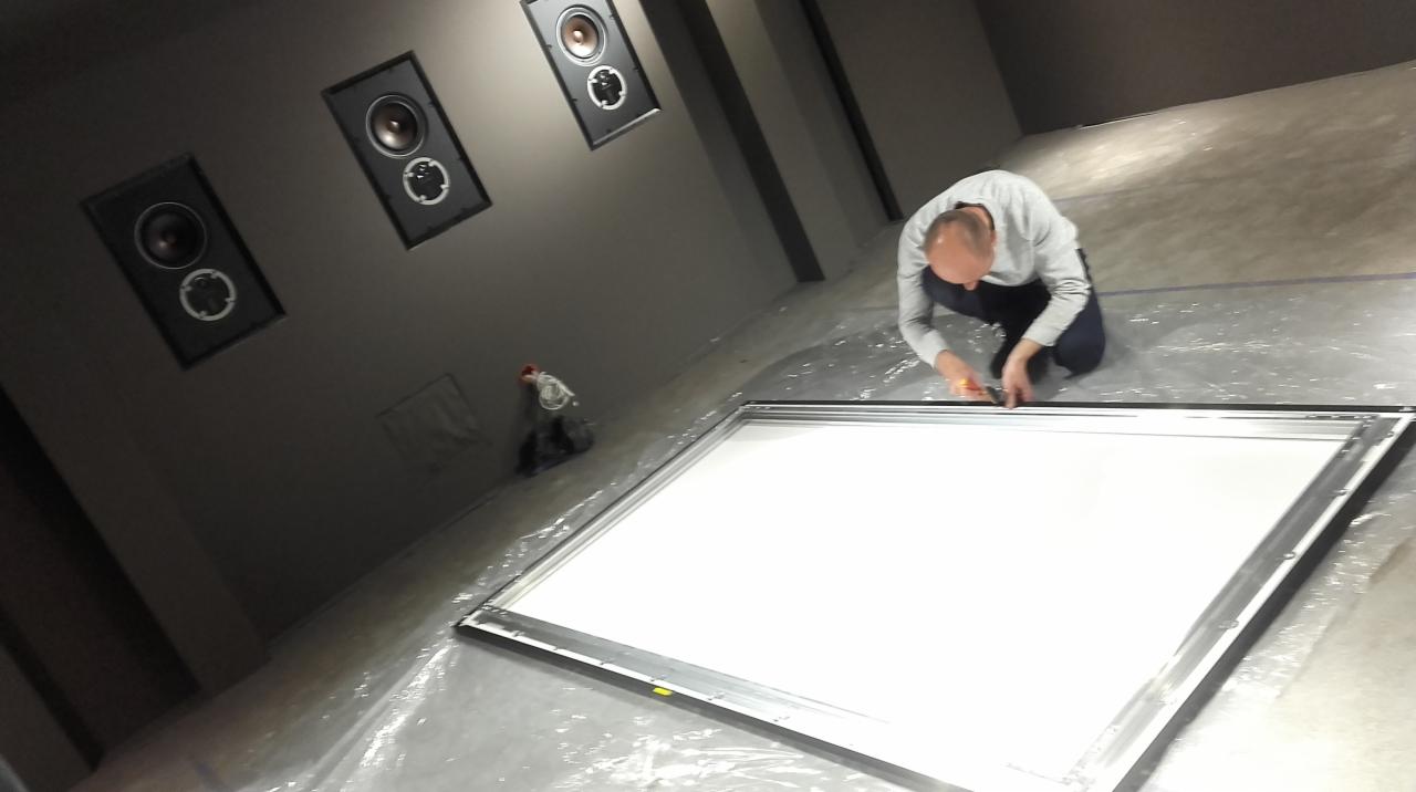 Instalacja ekranu projekcyjnego w pomieszczeniu z zamontowanymi głośnikami na ścianie we Wrześni. Pracownik w trakcie montażu ramy ekranu.